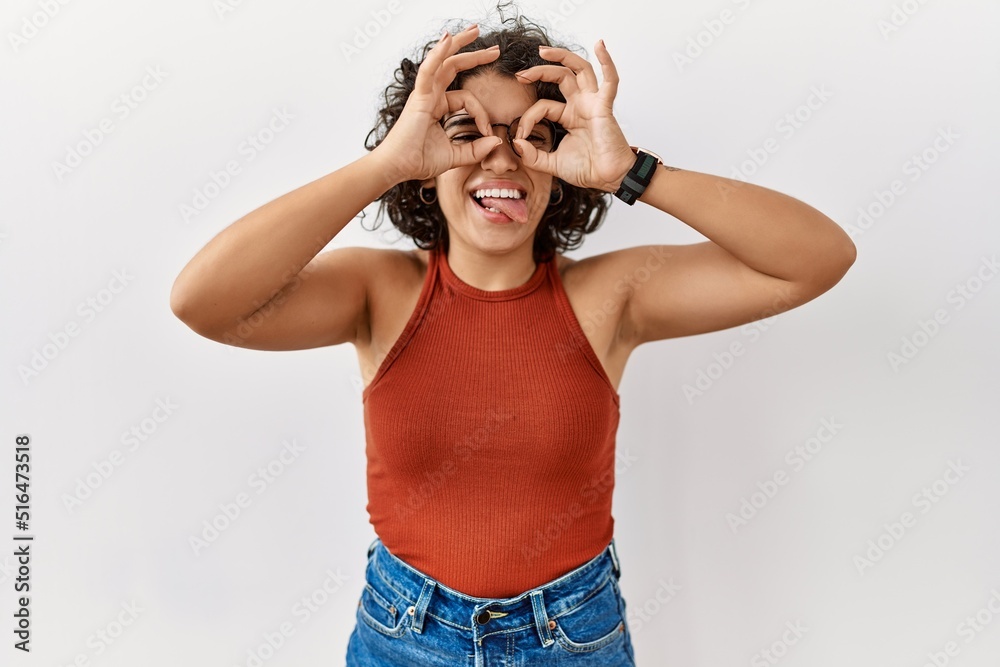 Young hispanic woman wearing glasses standing over isolated background doing ok gesture like binoculars sticking tongue out, eyes looking through fingers. crazy expression. Young hispanic woman wearing glasses standing over isolated background doing ok gesture like binoculars sticking tongue out, eyes looking through fingers. crazy expression.