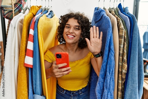 Young hispanic woman searching clothes on clothing rack using smartphone waiving saying hello happy and smiling, friendly welcome gesture