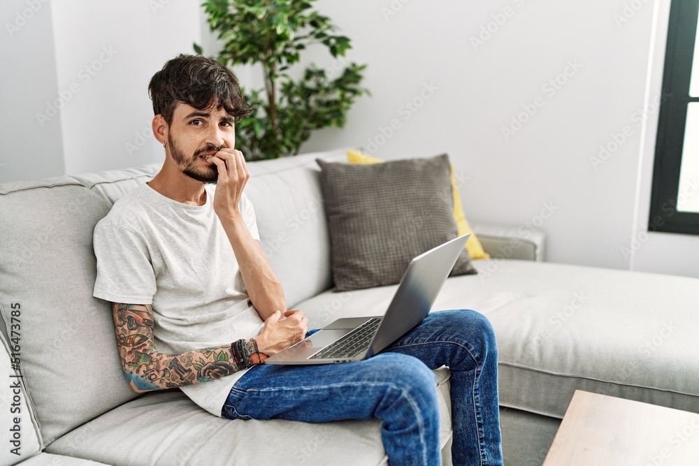 Hispanic man with beard sitting on the sofa looking stressed and nervous with hands on mouth biting nails. anxiety problem.