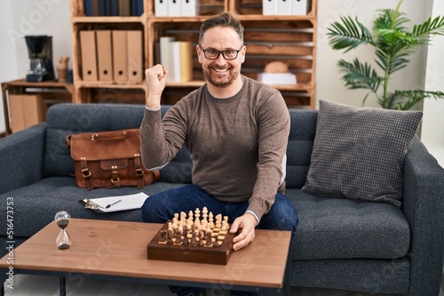 Middle age caucasian man playing chess sitting on the sofa screaming proud, celebrating victory and success very excited with raised arms
