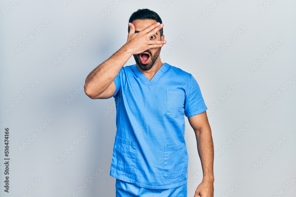 Handsome hispanic man with beard wearing blue male nurse uniform peeking in shock covering face and eyes with hand, looking through fingers with embarrassed expression. Handsome hispanic man with beard wearing blue male nurse uniform peeking in shock covering face and eyes with hand, looking through fingers with embarrassed expression.