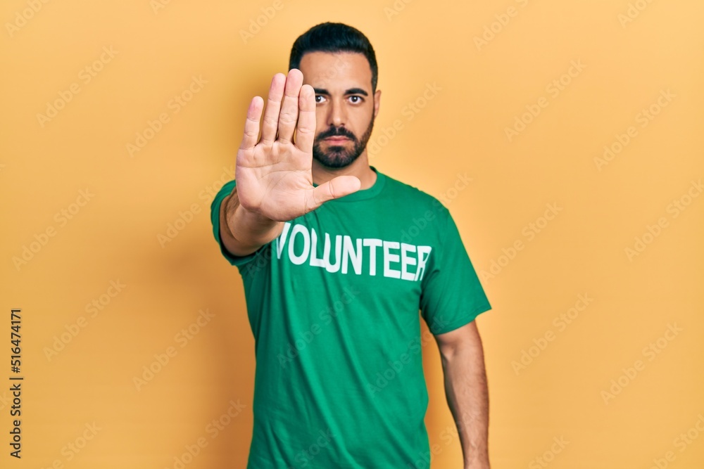 Handsome hispanic man with beard wearing volunteer t shirt with open hand doing stop sign with serious and confident expression, defense gesture Handsome hispanic man with beard wearing volunteer t shirt with open hand doing stop sign with serious and confident expression, defense gesture