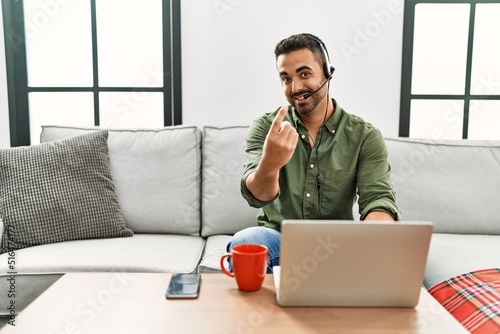 Young hispanic man with beard wearing call center agent headset working from home beckoning come here gesture with hand inviting welcoming happy and smiling