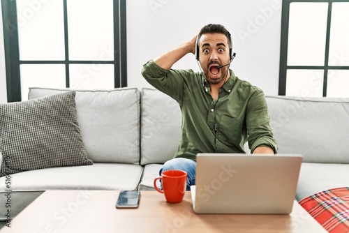 Young hispanic man with beard wearing call center agent headset working from home crazy and scared with hands on head, afraid and surprised of shock with open mouth