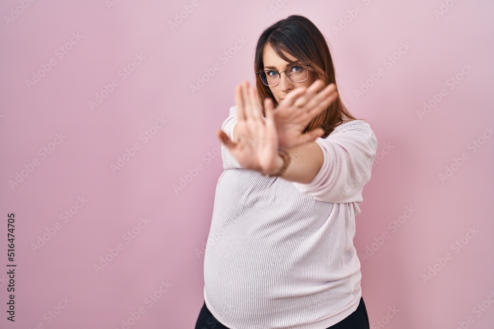 Pregnant woman standing over pink background rejection expression crossing arms and palms doing negative sign, angry face Pregnant woman standing over pink background rejection expression crossing arms and palms doing negative sign, angry face