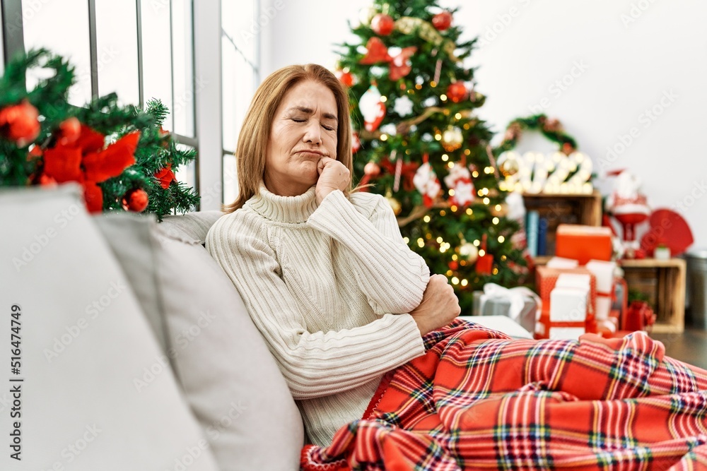 Middle age woman sitting on the sofa by christmas tree looking stressed and nervous with hands on mouth biting nails. anxiety problem.