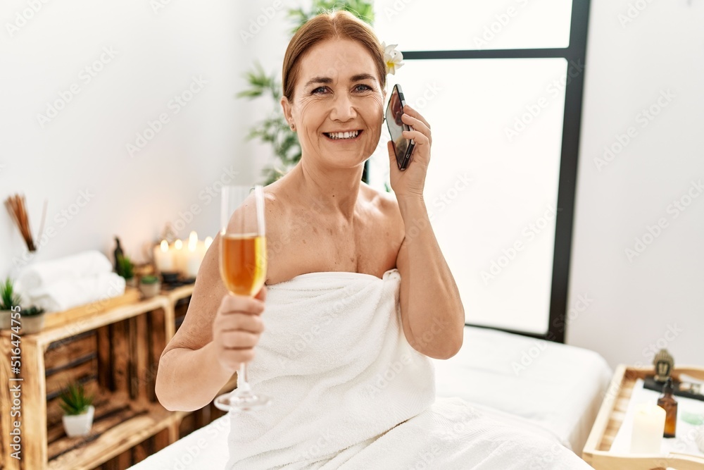 Middle age caucasian woman wearing towel talking on the smartphone sitting on massage board at beauty center Middle age caucasian woman wearing towel talking on the smartphone sitting on massage board at beauty center