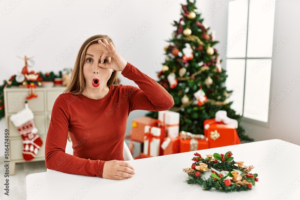 Caucasian young blonde woman sitting on the table by christmas tree doing ok gesture shocked with surprised face, eye looking through fingers. unbelieving expression. Caucasian young blonde woman sitting on the table by christmas tree doing ok gesture shocked with surprised face, eye looking through fingers. unbelieving expression.