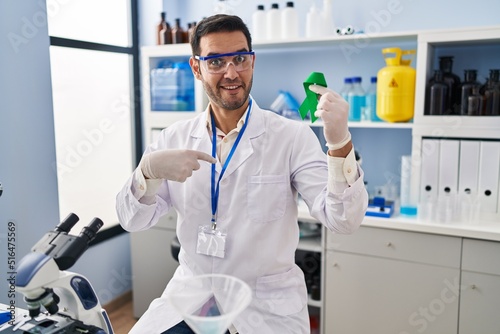 Young hispanic man with beard working at scientist laboratory holding green ribbon pointing finger to one self smiling happy and proud