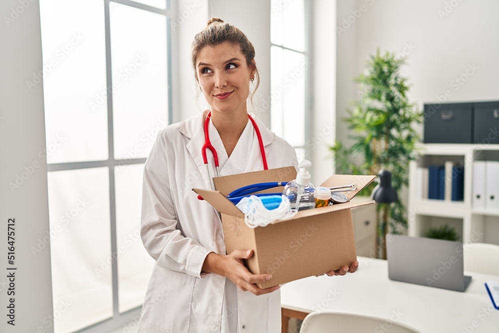 Young doctor woman holding box with medical items smiling looking to the side and staring away thinking.
