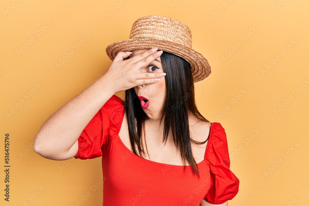 Young hispanic woman wearing summer hat peeking in shock covering face and eyes with hand, looking through fingers with embarrassed expression. Young hispanic woman wearing summer hat peeking in shock covering face and eyes with hand, looking through fingers with embarrassed expression.
