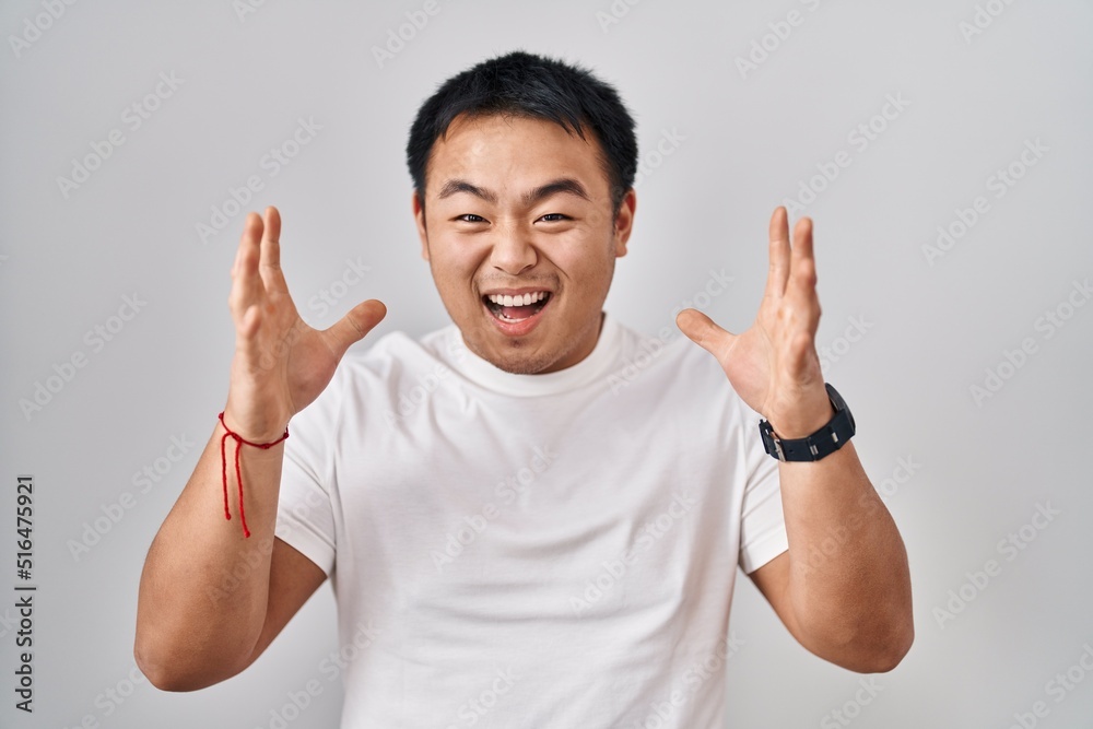 Young chinese man standing over white background celebrating mad and crazy for success with arms raised and closed eyes screaming excited. winner concept Young chinese man standing over white background celebrating mad and crazy for success with arms raised and closed eyes screaming excited. winner concept