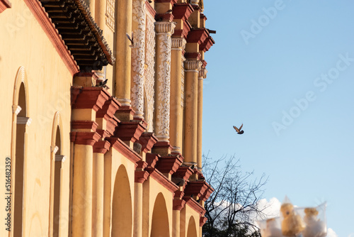 Yellow cathedral with pigeons flying around.