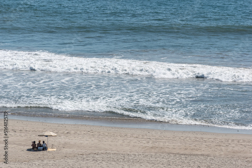 three people on the beach under an umbrella watching the ocean.