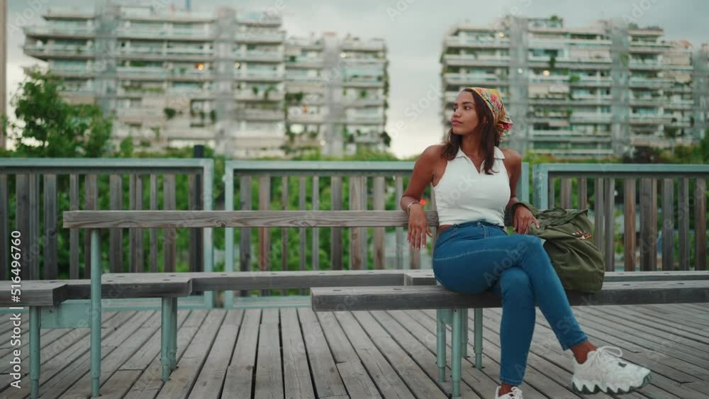 Cute tanned woman with long brown hair wearing white top and yellow bandana seated on bench on cityscape background. Camera moving forwards Cute tanned woman with long brown hair wearing white top and yellow bandana seated on bench on cityscape background. Camera moving forwards