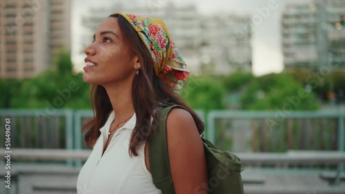 Clouse-up, cute tanned woman with long brown hair in white top and yellow bandana with backpack on her shoulder and looks around the modern city. Camera moving around photo