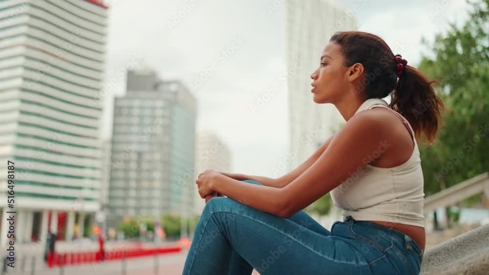 Profile of cute tanned woman with long brown hair wearing white top sits and looks thoughtfully at modern houses