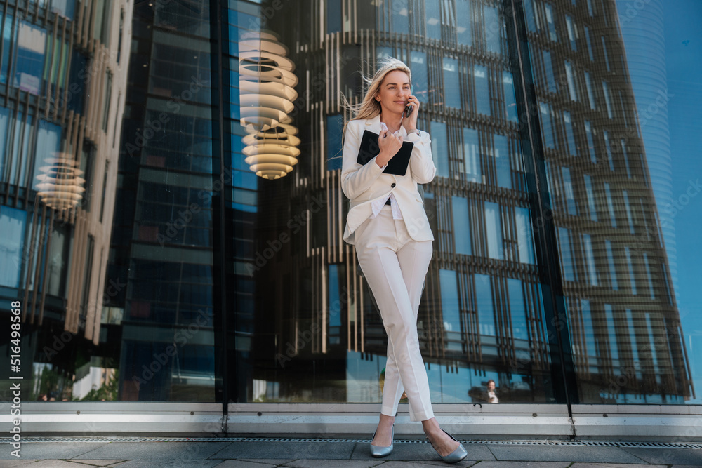 Elegant blonde young businesswoman in white suite standing outdoors against huge building with large windows. Satisfied talking by phone holding diary, satisfied by career. Business, finance concept.