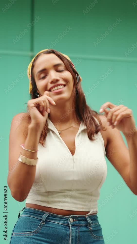 VERTICAL VIDEO: Cute tanned woman with long brown hair wearing white top and yellow bandana listens to music in wireless headphones and dances on blue wall background VERTICAL VIDEO: Cute tanned woman with long brown hair wearing white top and yellow bandana listens to music in wireless headphones and dances on blue wall background