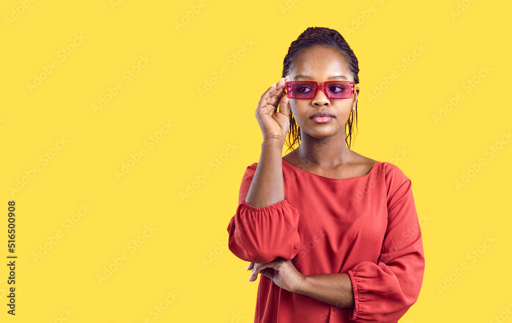 Fototapeta premium Portrait of cool and stylish young african american woman in red glasses on yellow background. Serious woman holding glasses on her face standing near copy space. Banner for website design.
