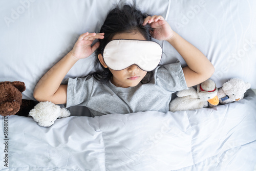 Top view child little asian girl blindfold sleeps in the bed with a toy teddy bear