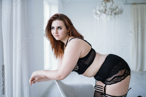 A plus size model in lacy black lingerie poses in a bright room with a trendy interior. Seductive chubby model in lingerie and harness