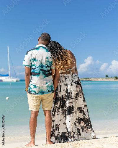 Young black couple stand on the beach while looking towards the horizon leaning heads on shoulders. Jamaican couple. Photographed from behind