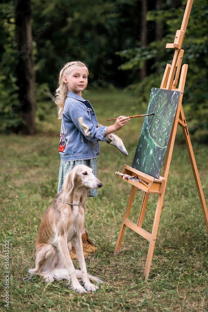girl stands in front of an easel with a dog draws