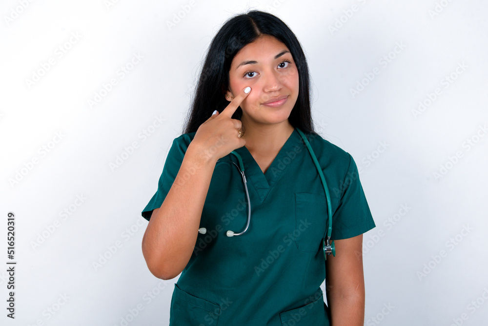 Doctor hispanic woman wearing surgeon uniform over white wall Pointing to the eye watching you gesture, suspicious expression. Doctor hispanic woman wearing surgeon uniform over white wall Pointing to the eye watching you gesture, suspicious expression.