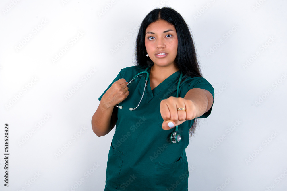 Doctor hispanic woman wearing surgeon uniform over white wall Punching fist to fight, aggressive and angry attack, threat and violence Doctor hispanic woman wearing surgeon uniform over white wall Punching fist to fight, aggressive and angry attack, threat and violence