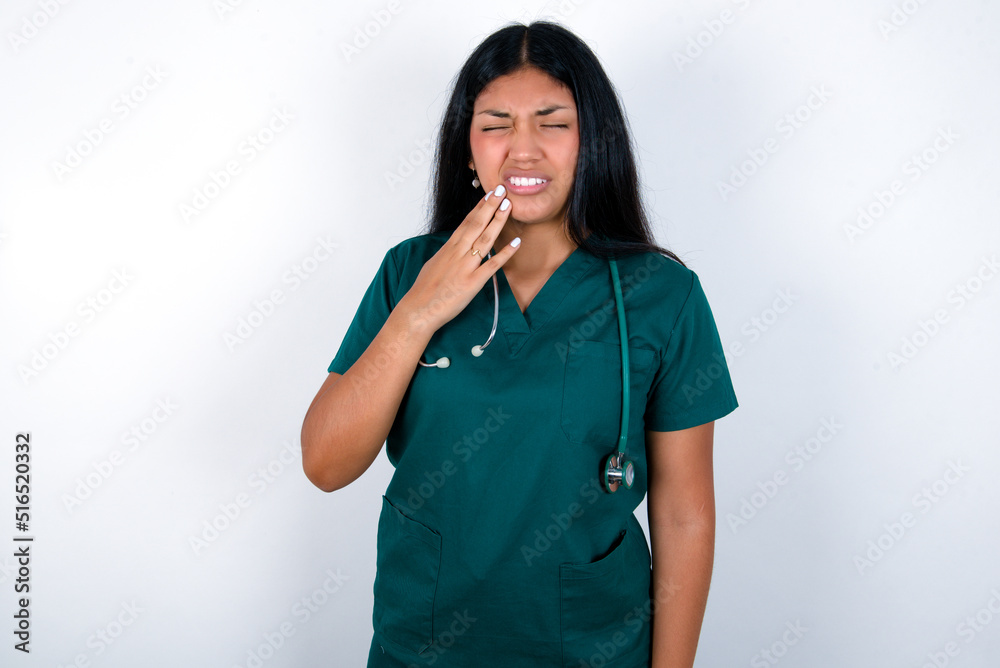 Doctor hispanic woman wearing surgeon uniform over white wall touching mouth with hand with painful expression because of toothache or dental illness on teeth. Doctor hispanic woman wearing surgeon uniform over white wall touching mouth with hand with painful expression because of toothache or dental illness on teeth.