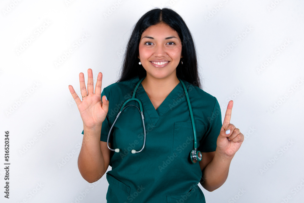 Doctor hispanic woman wearing surgeon uniform over white wall showing and pointing up with fingers number six while smiling confident and happy. Doctor hispanic woman wearing surgeon uniform over white wall showing and pointing up with fingers number six while smiling confident and happy.