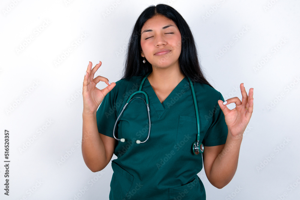 Doctor hispanic woman wearing surgeon uniform over white wall relax and smiling with eyes closed doing meditation gesture with fingers. Yoga concept. Doctor hispanic woman wearing surgeon uniform over white wall relax and smiling with eyes closed doing meditation gesture with fingers. Yoga concept.