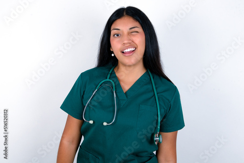 Doctor hispanic woman wearing surgeon uniform over white wall winking looking at the camera with sexy expression, cheerful and happy face.