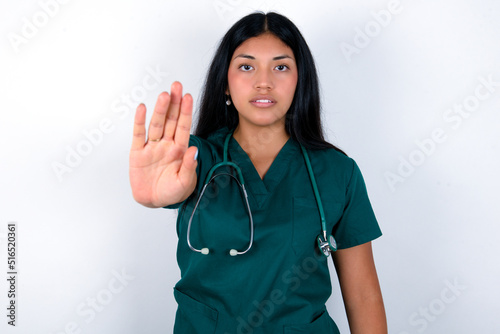 Doctor hispanic woman wearing surgeon uniform over white wall doing stop sing with palm of the hand. Warning expression with negative and serious gesture on the face.