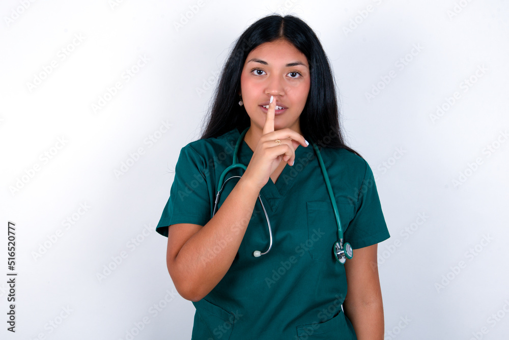 Doctor hispanic woman wearing surgeon uniform over white wall makes silence gesture, keeps finger over lips. Silence and secret concept. Doctor hispanic woman wearing surgeon uniform over white wall makes silence gesture, keeps finger over lips. Silence and secret concept.