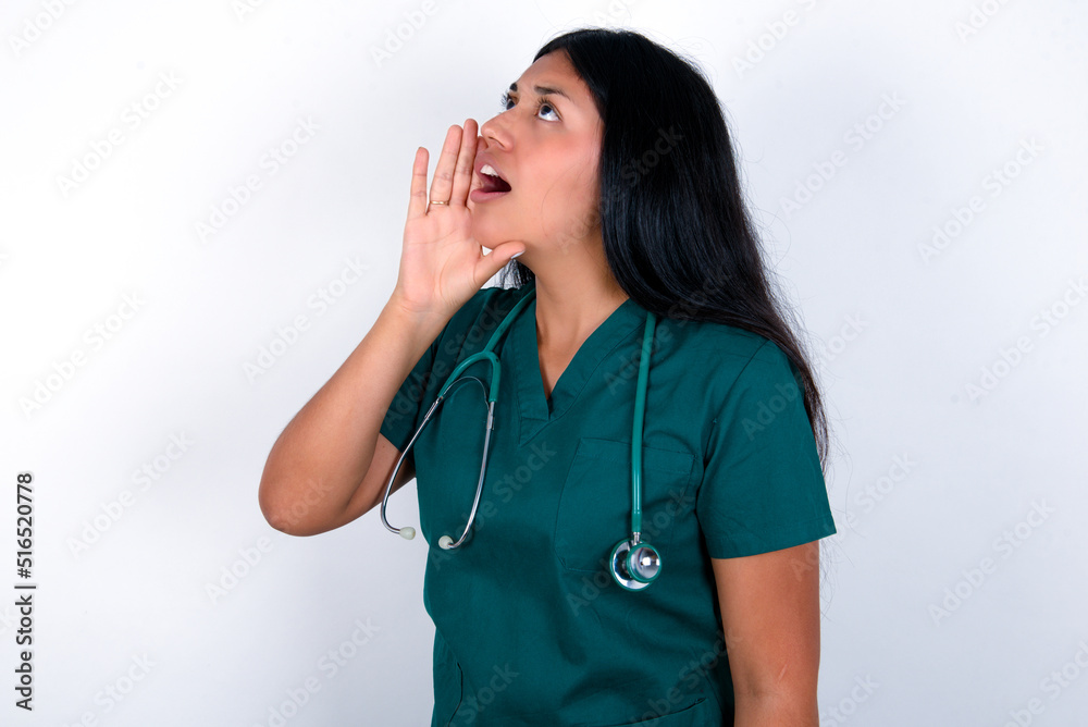 Doctor hispanic woman wearing surgeon uniform over white wall shouting and screaming loud to side with hand on mouth. Communication concept. Doctor hispanic woman wearing surgeon uniform over white wall shouting and screaming loud to side with hand on mouth. Communication concept.