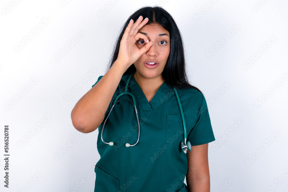Doctor hispanic woman wearing surgeon uniform over white wall doing ok gesture shocked with surprised face, eye looking through fingers. Unbelieving expression. Doctor hispanic woman wearing surgeon uniform over white wall doing ok gesture shocked with surprised face, eye looking through fingers. Unbelieving expression.