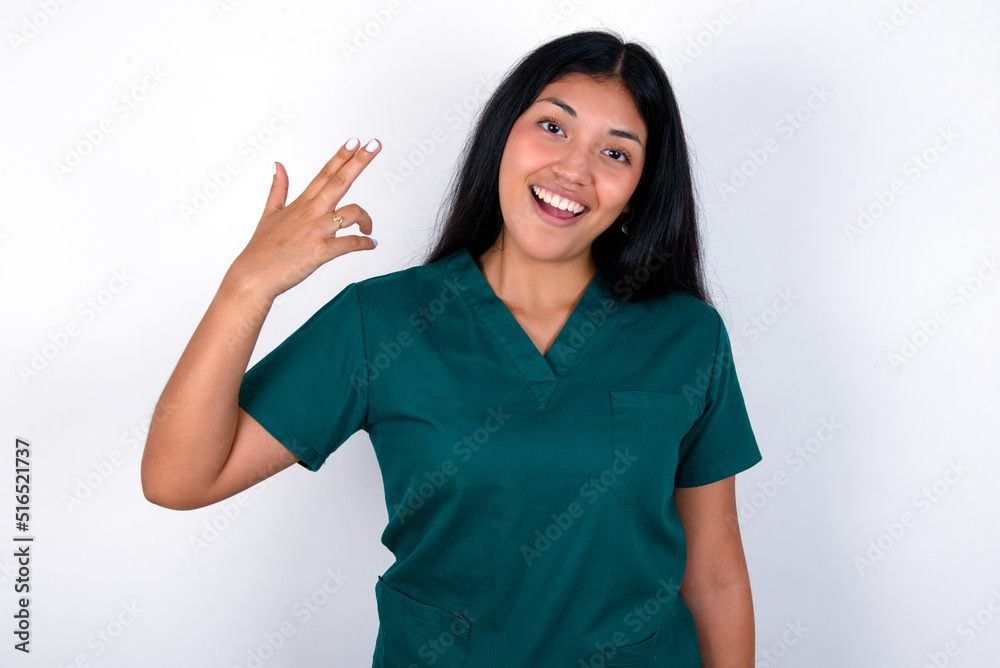 Doctor hispanic woman wearing surgeon uniform over white wall foolishness around shoots in temple with fingers makes suicide gesture. Funny model makes finger gun pistol Doctor hispanic woman wearing surgeon uniform over white wall foolishness around shoots in temple with fingers makes suicide gesture. Funny model makes finger gun pistol