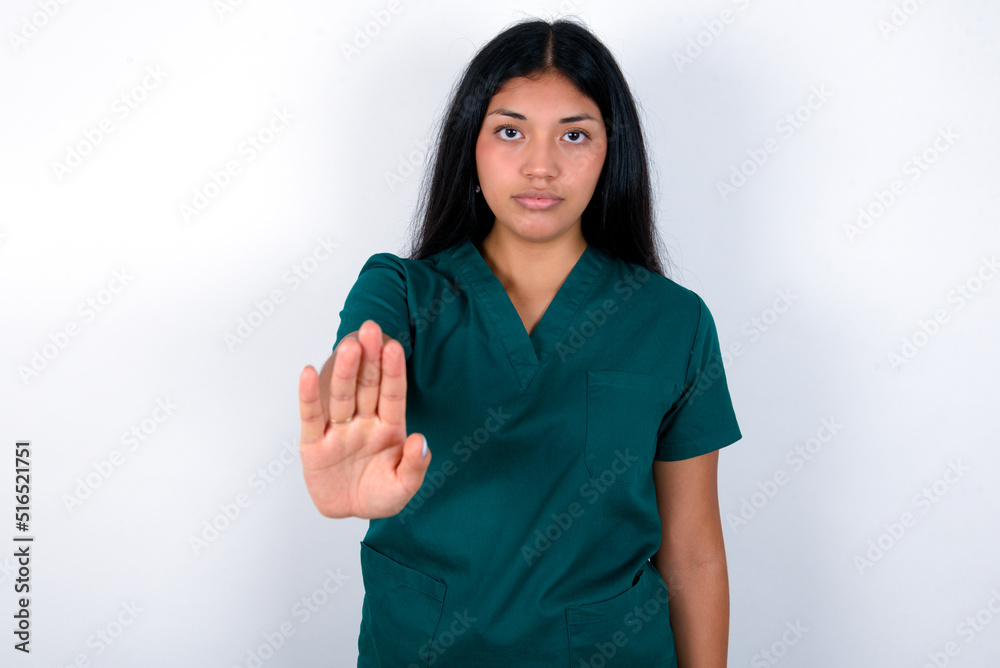 Doctor hispanic woman wearing surgeon uniform over white wall shows stop sign prohibition symbol keeps palm forward to camera with strict expression Doctor hispanic woman wearing surgeon uniform over white wall shows stop sign prohibition symbol keeps palm forward to camera with strict expression