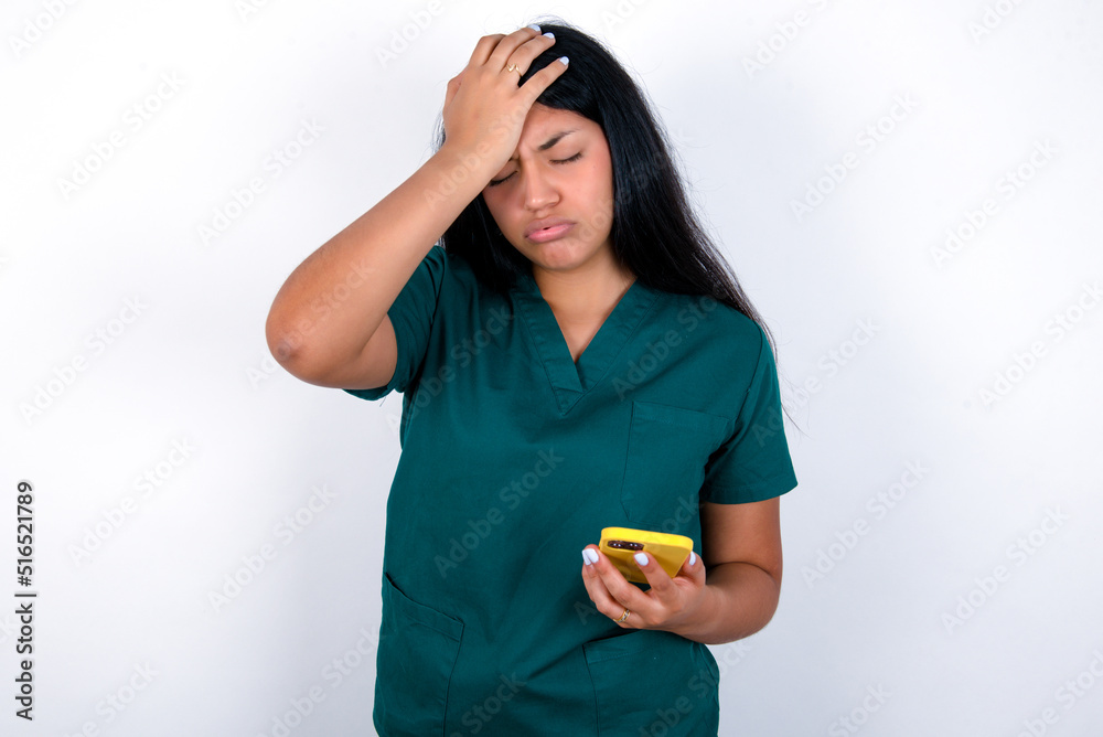Upset depressed Doctor hispanic woman wearing surgeon uniform over white background makes face palm as forgot about something important holds mobile phone expresses sorrow and regret blames Upset depressed Doctor hispanic woman wearing surgeon uniform over white background makes face palm as forgot about something important holds mobile phone expresses sorrow and regret blames