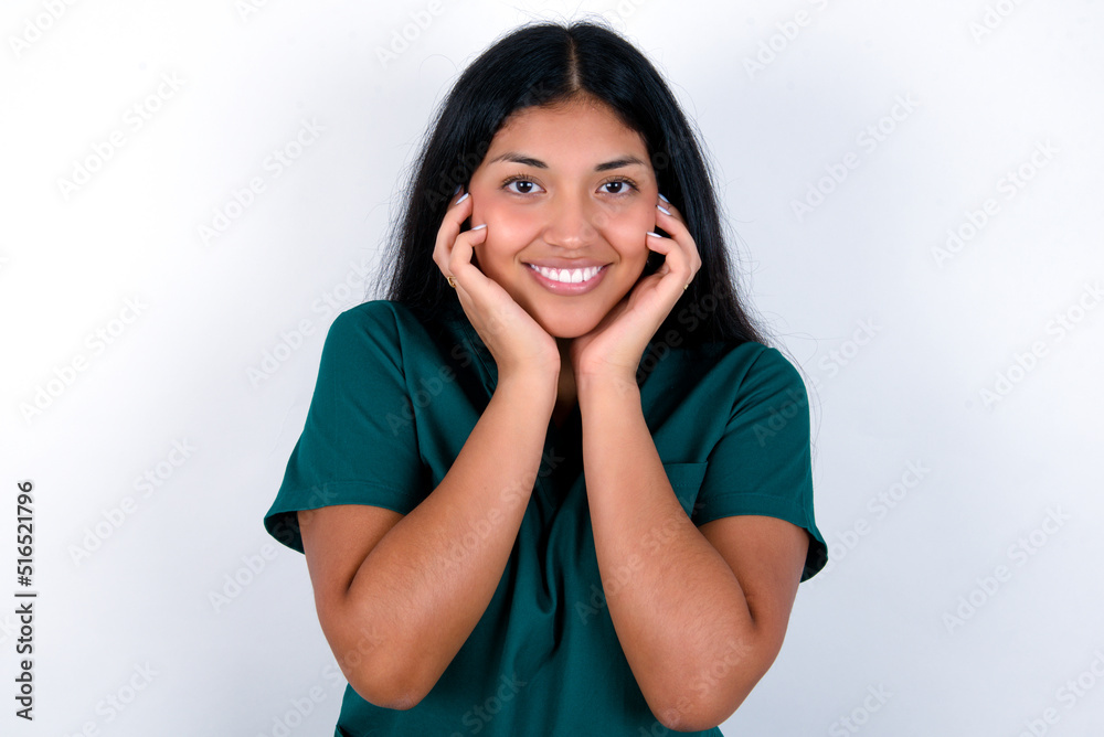 Happy Doctor hispanic woman wearing surgeon uniform over white background touches both cheeks gently, has tender smile, shows white teeth, gazes positively straightly at camera, Happy Doctor hispanic woman wearing surgeon uniform over white background touches both cheeks gently, has tender smile, shows white teeth, gazes positively straightly at camera,