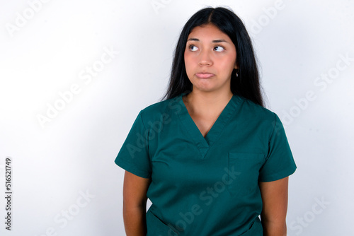 Dissatisfied Doctor hispanic woman wearing surgeon uniform over white wall purses lips and has unhappy expression looks away stands offended. Depressed frustrated model. © Roquillo
