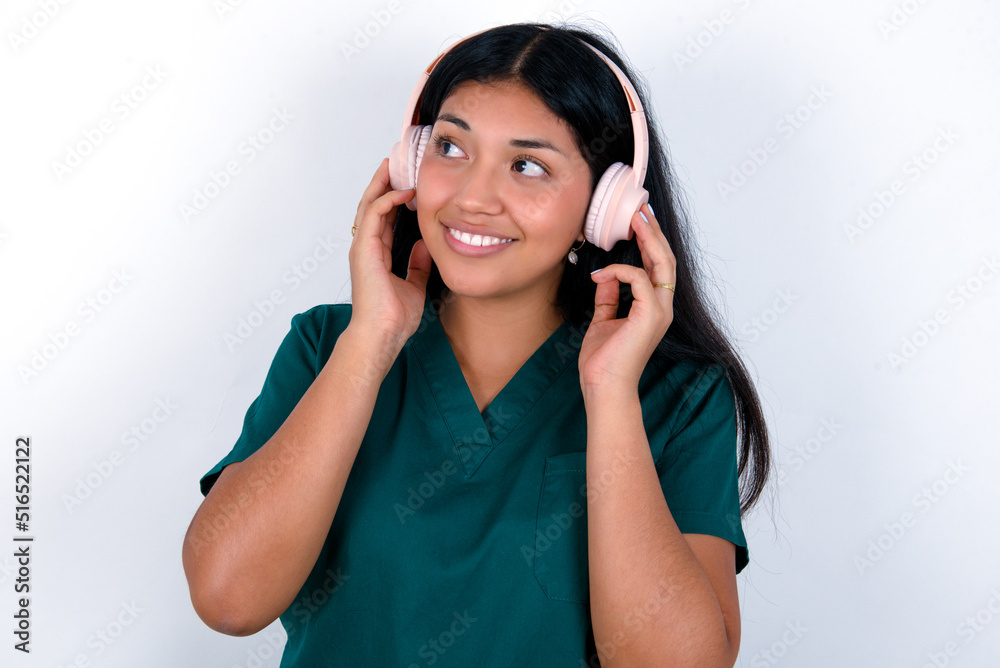 Doctor hispanic woman wearing surgeon uniform over white background wears stereo headphones listens music concentrated aside. People hobby lifestyle concept Doctor hispanic woman wearing surgeon uniform over white background wears stereo headphones listens music concentrated aside. People hobby lifestyle concept