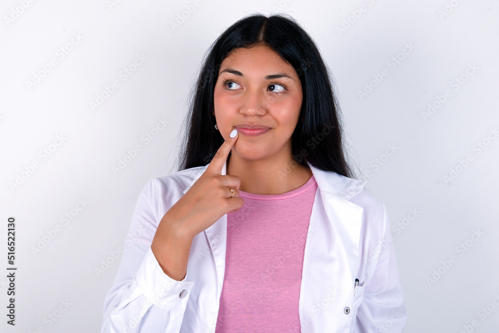 Lovely dreamy Young hispanic doctor girl wearing coat over white background keeps finger near lips looks aside copy space. Lovely dreamy Young hispanic doctor girl wearing coat over white background keeps finger near lips looks aside copy space.