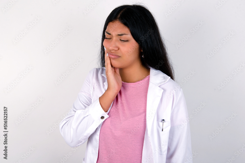 Young hispanic doctor girl wearing coat over white background with toothache Young hispanic doctor girl wearing coat over white background with toothache