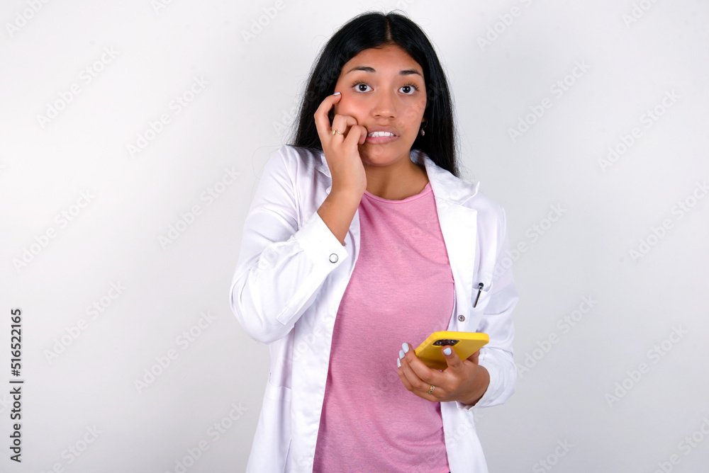 Afraid funny Young hispanic doctor girl wearing coat over white background holding telephone and bitting nails Afraid funny Young hispanic doctor girl wearing coat over white background holding telephone and bitting nails