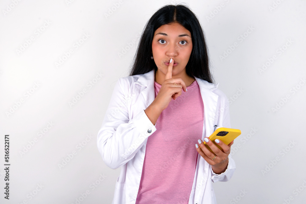 Young hispanic doctor girl wearing coat over white background holding modern gadget ask not tell secrets Young hispanic doctor girl wearing coat over white background holding modern gadget ask not tell secrets