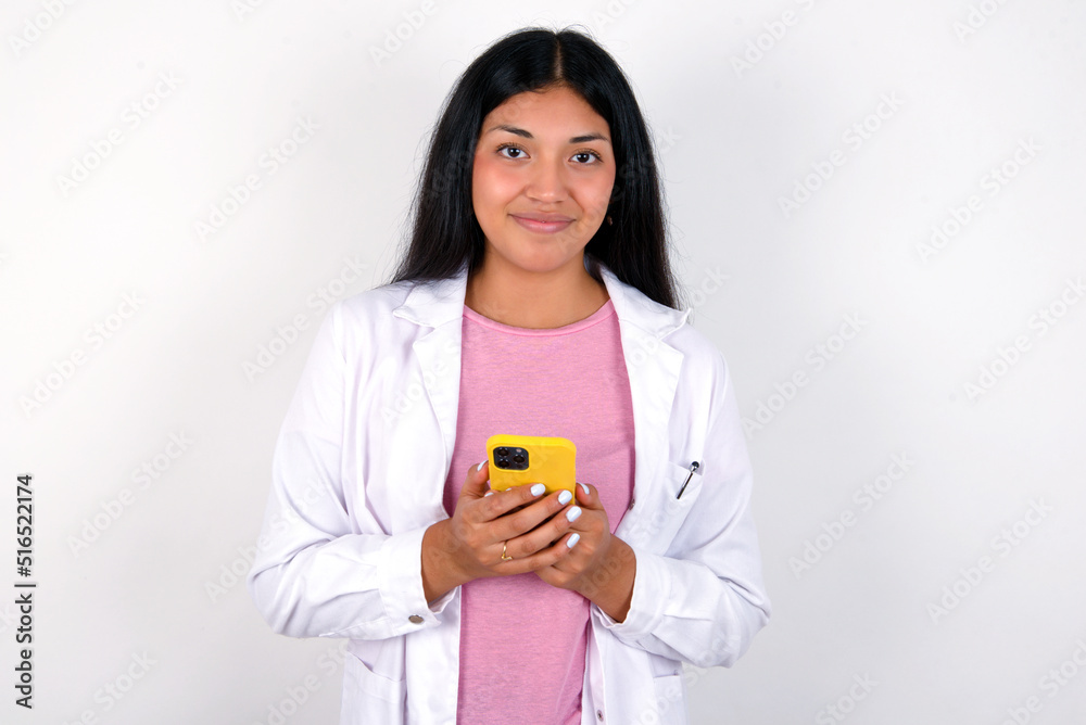 Portrait of serious confident Young hispanic doctor girl wearing coat over white background holding phone in two hands Portrait of serious confident Young hispanic doctor girl wearing coat over white background holding phone in two hands