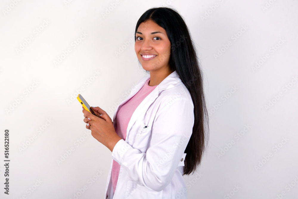 Rear view photo portrait of Young hispanic doctor girl wearing coat over white background using smartphone smiling Rear view photo portrait of Young hispanic doctor girl wearing coat over white background using smartphone smiling